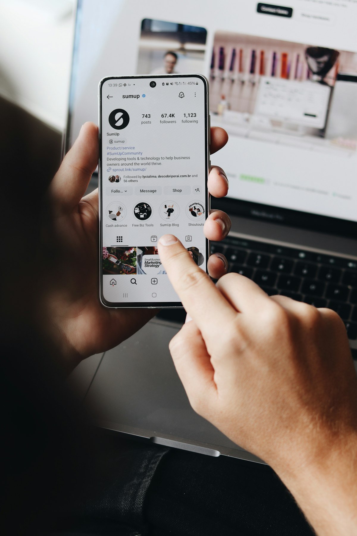 Hand holding a phone with a social media feed in front of a laptop